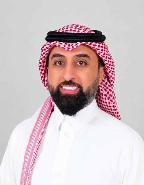 رجل ملتحٍ يرتدي الثوب الأبيض والشماغ الأحمر والعقال الأسود، يبتسم بوضوح أمام خلفية رمادية.